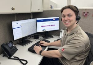 A 211 Call Specialist, headset in place, seated at their computer while facing the camera. A 211 Call Specialist, headset in place, seated at their computer while facing the camera.
